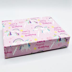 Puede incluir: Papel de regalo rosa con un patrón de unicornio y arcoíris. El papel tiene el texto "Have a Magical Birthday" repetido en toda su superficie.