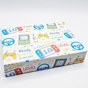 May include: White wrapping paper with a repeated pattern of video game controllers, steering wheels, and game consoles. The paper features the text "Ready On Your Birthday For The Next Level" and "Level Up".