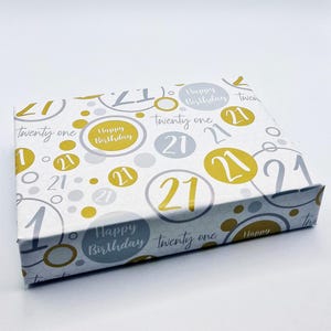 Puede incluir: Una caja de regalo blanca con un patrón dorado y plateado. La caja está decorada con el número 21 y las palabras "Feliz cumpleaños" y "Veintiún".