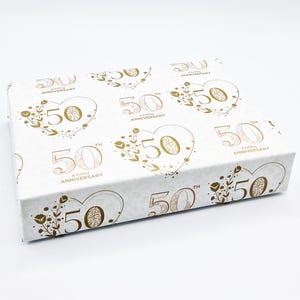 May include: White gift box with a gold pattern of hearts and the text "50th Anniversary" repeated. The box is decorated with gold floral accents.