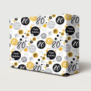 Puede incluir: Una caja de regalo envuelta en papel blanco con un patrón repetido de círculos negros, dorados y grises. Cada círculo contiene el número "80" o las palabras "Happy Birthday" y "eighty".