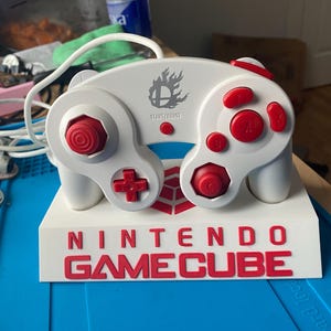 Puede incluir: Un mando blanco de Nintendo GameCube con botones y detalles en rojo, incluyendo la cruceta y los joysticks analógicos. El mando descansa sobre una base blanca con las palabras "NINTENDO GAME CUBE" en rojo. El mando tiene el logo de Super Smash Bros.