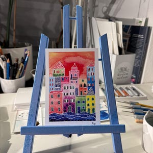 May include: A watercolour painting of a colourful cityscape with a red, orange and pink sunset in the background. The buildings are painted in a variety of bright colours, including yellow, green, blue and purple. The painting is signed "Ro" in the bottom right corner.