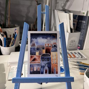 May include: A watercolor painting of a small town with colorful houses and a crescent moon in the night sky. The painting is on a blue easel.