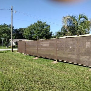 May include: A brown privacy fence made of mesh fabric is installed in a yard. The fence is secured to wooden posts and is about 6 feet tall.