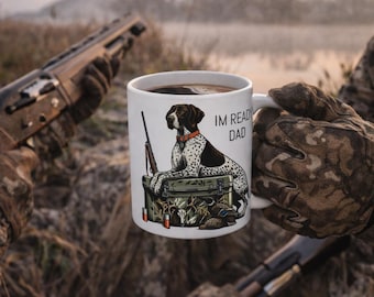 Könnte beinhalten: Weiße Keramiktasse mit einer Jagdhunde-Illustration, Gewehr und dem Text "IM READY DAD". Der Hund ist schwarz-weiß mit rotem Halsband. Die Tasse wird von einer Person mit Camouflage-Handschuhen gehalten.