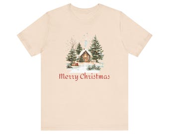 Puede incluir: Camiseta color crema con una ilustración en acuarela de una cabaña cubierta de nieve rodeada de árboles de hoja perenne. La frase "Merry Christmas" está impresa en rojo debajo de la imagen, evocando un tema festivo navideño.