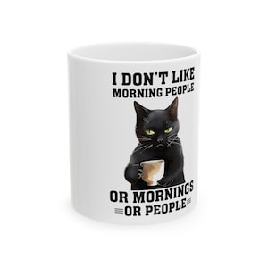 Op de afbeelding: Witte keramische mok met de tekst "I DON'T LIKE MORNING PEOPLE OR MORNINGS OR PEOPLE." Een zwarte kat met gele ogen houdt een koffiekopje vast. De mok heeft een standaard formaat en vorm.