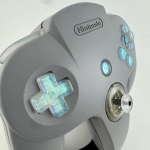 Puede incluir: Un mando gris de Nintendo 64 con un joystick transparente y botones iridiscentes. El mando presenta una cruceta direccional y el logotipo de Nintendo. Los botones tienen un efecto brillante y holográfico.