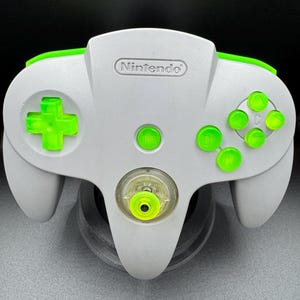 May include: A gray Nintendo 64 video game controller featuring bright neon green buttons, a directional pad, and a central analog stick. The controller includes a start button and four C-buttons, all in a matching neon green translucent finish.
