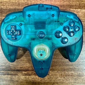 May include: A translucent teal Nintendo 64 video game controller featuring a central analog stick, a directional pad on the left, A and B buttons, four C-buttons on the right, and a central start button. The internal circuitry is visible through the shell.