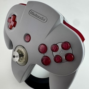 Puede incluir: Un mando gris de Nintendo 64 con detalles en rojo. El mando presenta una cruceta, un botón de inicio y botones C rojos. El logotipo de Nintendo es visible en la parte superior.