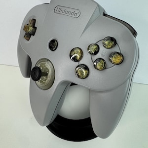 Puede incluir: Un mando gris de Nintendo 64 con botones dorados y un joystick. El logotipo de Nintendo está en la parte superior. Un accesorio de juego clásico.