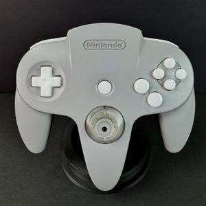 May include: A gray Nintendo 64 controller with white buttons and a directional pad. The controller has a clear, circular center piece and the Nintendo logo at the top. It is displayed on a black stand.