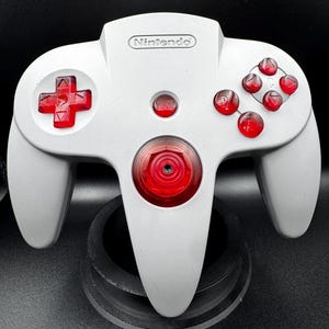 Peut inclure: Une manette de jeu vidéo Nintendo 64 gris clair avec des boutons rouge translucide, une croix directionnelle rouge et un stick analogique central rouge. Le mot Nintendo est gravé en haut au centre, avec un bouton Start rouge au-dessus du stick.