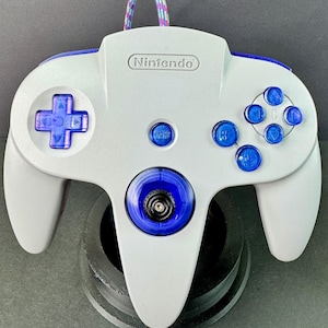 Puede incluir: Un mando gris de Nintendo 64 con detalles en azul y morado. El mando tiene una cruceta, botones y un joystick. El logotipo de Nintendo es visible. Un cable colorido está conectado.