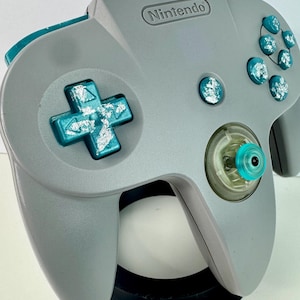Puede incluir: Un mando gris de Nintendo 64 con detalles turquesa. El mando tiene una cruceta, botones y una palanca analógica. La palabra "Nintendo" está impresa en la parte superior.
