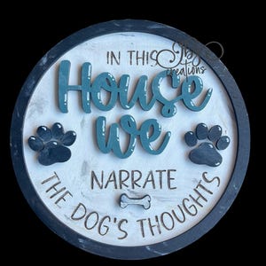 May include: A round wooden sign with a black border and a white background. The sign says "In this house we narrate the dog's thoughts" with a bone icon and two paw prints. The words "house" and "we" are in teal blue.