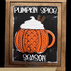 May include: A wooden framed sign with a black chalkboard background. The sign features a pumpkin spice latte with whipped cream and cinnamon sticks. The text "Pumpkin Spice Season" is written in white on the chalkboard.