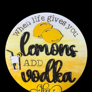 May include: A round wooden sign with a yellow and white ombre background. The sign says "When life gives you lemons add vodka" in black lettering. There are two yellow lemons and a black silhouette of a glass with a straw and a lemon slice.