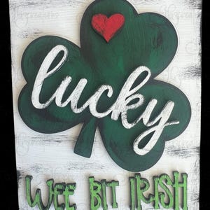May include: A wooden sign with a green shamrock and the words "lucky wee bit irish" in white and green lettering. A red heart is in the center of the shamrock.