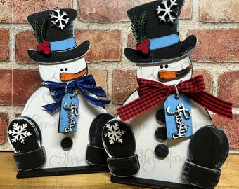 May include: Two wooden snowman figurines with black top hats, blue scarves, and red and black plaid bows. Each snowman has a black button nose, a black and white snowflake on their mittens, and a blue tag that says "Frosty".