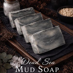 Puede incluir: Cuatro barras rectangulares de jabón de barro del Mar Muerto con un patrón veteado gris y blanco están dispuestas sobre una bandeja de madera sobre una tela negra. Las palabras "Dead Sea Mud Soap" y "100% Natural" están impresas.