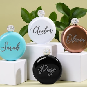Pode incluir: Quatro frascos redondos personalizados com rolhas adornadas com joias. Os frascos são turquesa, branco, cobre e preto. Os nomes "Sarah", "Amber", "Olivia" e "Page" estão gravados nos frascos.