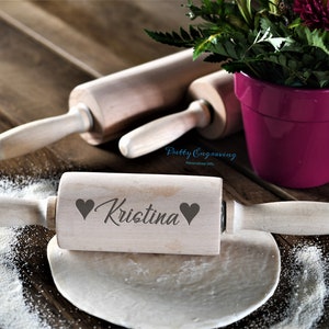 K&ouml;nnte beinhalten: Ein Holz-Nudelholz mit dem Namen "Kristina" eingraviert, umgeben von zwei Herzen. Das Nudelholz liegt auf einer Holzoberfl&auml;che mit etwas verstreuter Mehl.