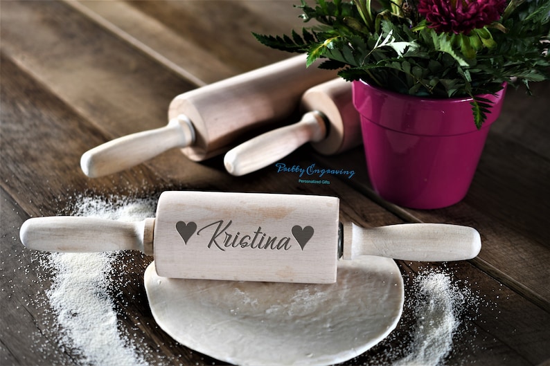 K&ouml;nnte beinhalten: Ein Holz-Nudelholz mit dem Namen "Kristina" eingraviert, umgeben von zwei Herzen. Das Nudelholz liegt auf einer Holzoberfl&auml;che mit etwas verstreuter Mehl.