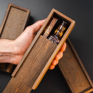 May include: A wooden box with a lid open, revealing a small bottle of liquor and a cigar. The box is made of dark wood with a natural grain.