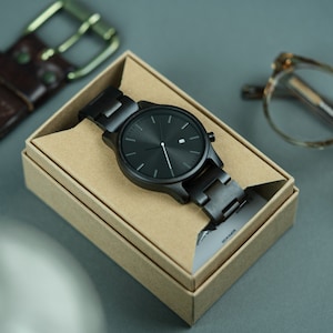 May include: A black wristwatch with a black metal band in a brown cardboard box. The watch has a black face with silver hands and a date window.