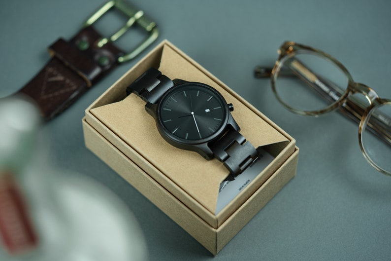 May include: A black wristwatch with a black metal band in a brown cardboard box. The watch has a black face with silver hands and a date window.
