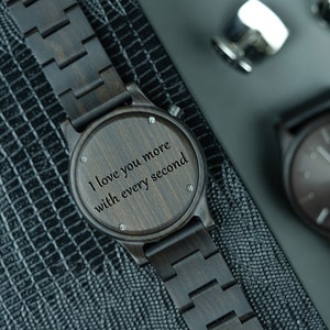 May include: A dark brown wooden wristwatch with a black band. The watch face has a wood grain pattern and the words "I love you more with every second" engraved on it.