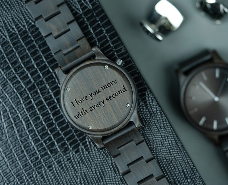 May include: A dark brown wooden wristwatch with a black band. The watch face has a wood grain pattern and the words "I love you more with every second" engraved on it.