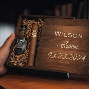 May include: A wooden box with a personalized inscription for a groom. The box contains a small bottle of whiskey and a cigar.