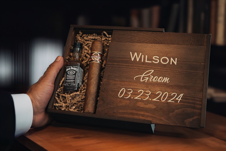 May include: A wooden box with a personalized inscription for a groom. The box contains a small bottle of whiskey and a cigar.