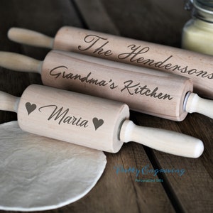 K&ouml;nnte beinhalten: Drei Holz-Nudelh&ouml;lzer mit personalisierten Gravuren. Das obere Nudelholz sagt "The Hendersons", das mittlere Nudelholz sagt "Grandma's Kitchen" und das untere Nudelholz sagt "Maria" mit zwei Herzen.