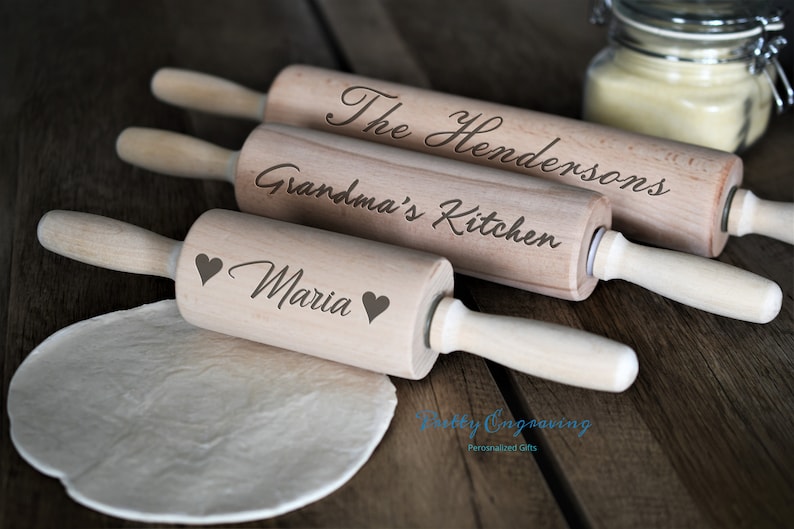 K&ouml;nnte beinhalten: Drei Holz-Nudelh&ouml;lzer mit personalisierten Gravuren. Das obere Nudelholz sagt "The Hendersons", das mittlere Nudelholz sagt "Grandma's Kitchen" und das untere Nudelholz sagt "Maria" mit zwei Herzen.