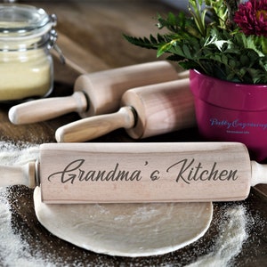 K&ouml;nnte beinhalten: Ein Holz-Nudelholz mit dem eingravierten Text "Grandma's Kitchen". Das Nudelholz liegt auf einem Holztisch mit einem Teigball und einem Glas Mehl.