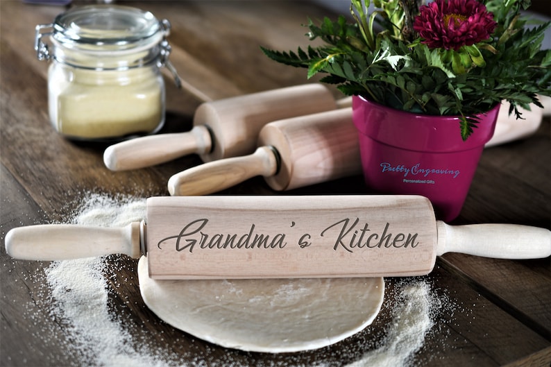 K&ouml;nnte beinhalten: Ein Holz-Nudelholz mit dem eingravierten Text "Grandma's Kitchen". Das Nudelholz liegt auf einem Holztisch mit einem Teigball und einem Glas Mehl.