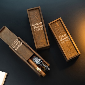 May include: Three wooden boxes with engraved names and dates. The boxes are closed except for one, which is open to reveal a small bottle of liquor and a cigar.
