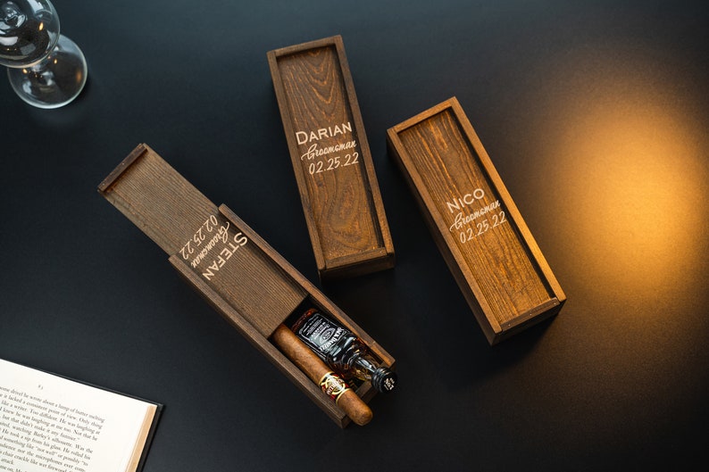 May include: Three wooden boxes with engraved names and dates. The boxes are closed except for one, which is open to reveal a small bottle of liquor and a cigar.