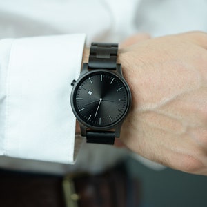 May include: A black wristwatch with a wooden band. The watch face is black with white hands and a small white square at the 12 o'clock position.