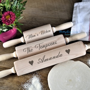 K&ouml;nnte beinhalten: Drei Holz-Nudelh&ouml;lzer mit personalisierten Gravuren. Das obere Nudelholz tr&auml;gt die Aufschrift "Mom's Kitchen", das mittlere Nudelholz tr&auml;gt die Aufschrift "The Tompsons" und das untere Nudelholz tr&auml;gt die Aufschrift "Amanda" mit einem Herzsymbol.