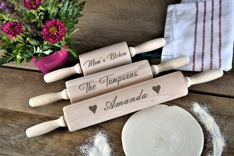 K&ouml;nnte beinhalten: Drei Holz-Nudelh&ouml;lzer mit personalisierten Gravuren. Das obere Nudelholz tr&auml;gt die Aufschrift "Mom's Kitchen", das mittlere Nudelholz tr&auml;gt die Aufschrift "The Tompsons" und das untere Nudelholz tr&auml;gt die Aufschrift "Amanda" mit einem Herzsymbol.