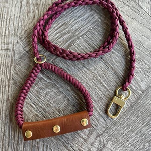 Puede incluir: Una correa trenzada burdeos para perros con un asa de cuero marrón asegurada con tres remaches dorados. La correa tiene un cierre dorado y un anillo dorado que conecta el asa con la cuerda trenzada.