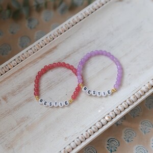 Könnte beinhalten: Zwei Perlenarmbänder mit dem Wort "BESTIE" in weißen Buchstaben auf jedem Armband. Das Armband links ist rosa und das Armband rechts ist lila.