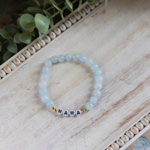 Könnte beinhalten: Ein hellblaues Perlenarmband mit dem Wort "MAMA" in goldenen Buchstaben.