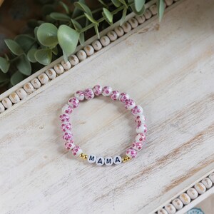 Könnte beinhalten: Weißes und rosafarbenes Blumenarmband mit goldenen Perlen und dem Wort "MAMA" in goldenen Buchstaben.